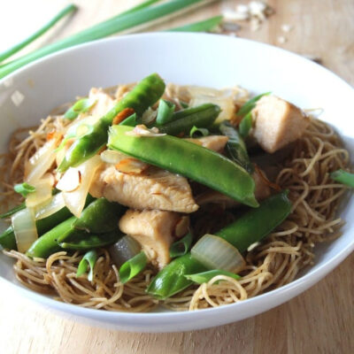 A bowl of Chinese Almond Chicken topped with fresh green snap peas on a wooden table.