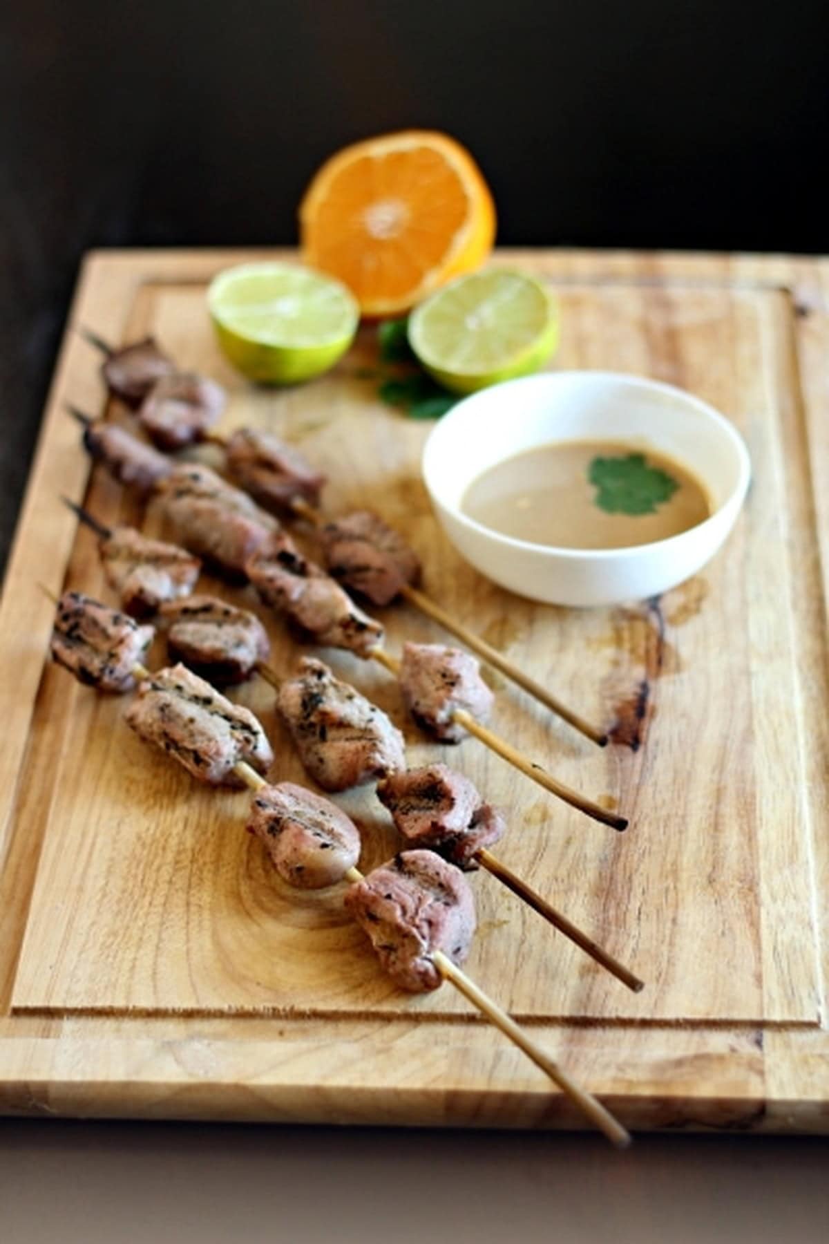 Multiple Indonesian Pork Satay skewers arranged on a wooden board with lime and orange halves.