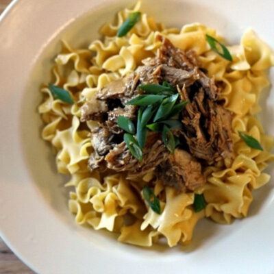 A plate of Pork Noodles topped with shredded meat and garnished with green herbs.