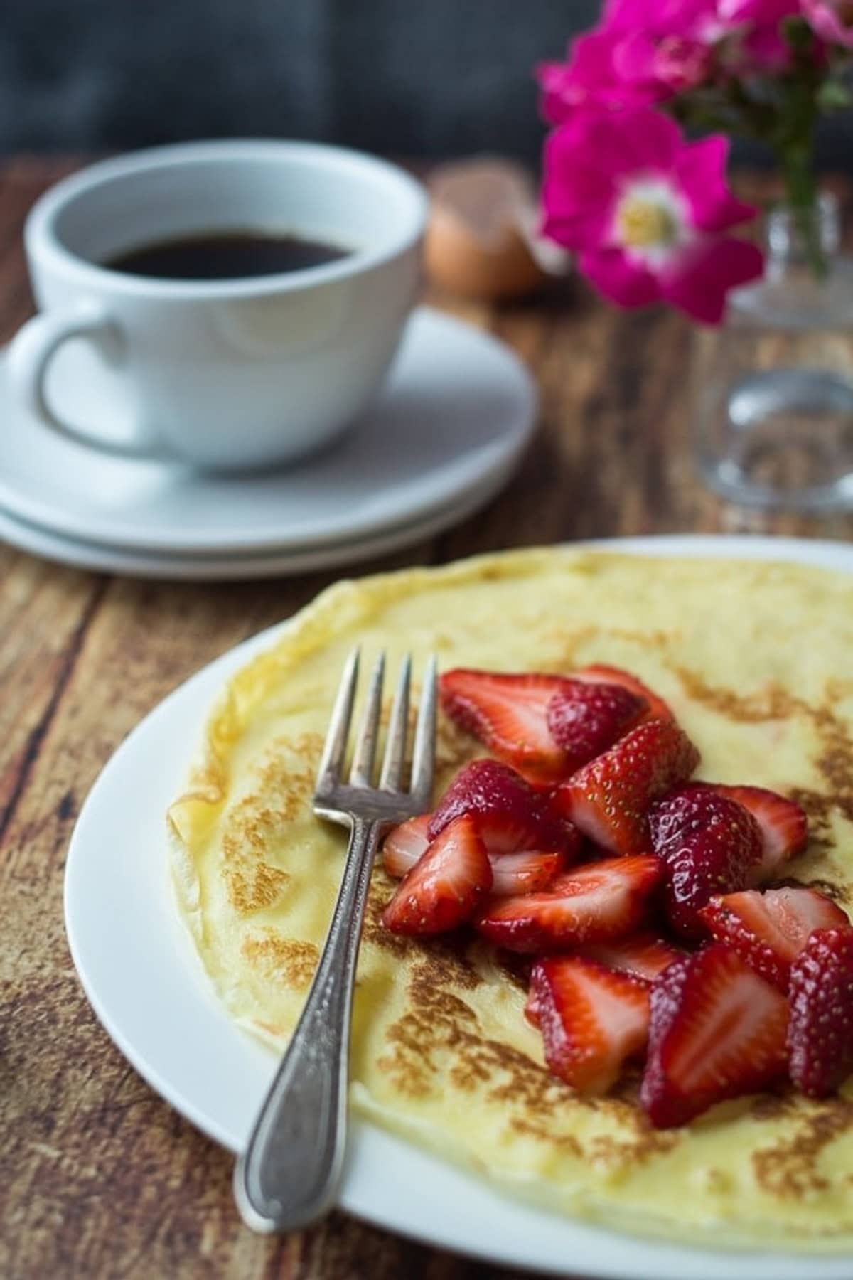 A plate of Swedish pancakes topped with fresh strawberries, next to a cup of black coffee.