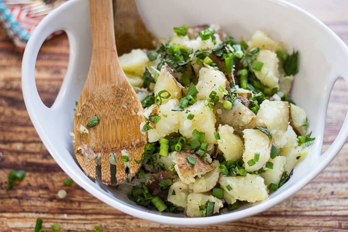 A bowl of Lebanese potato salad with chopped green onions and herbs, mixed with a wooden spoon.