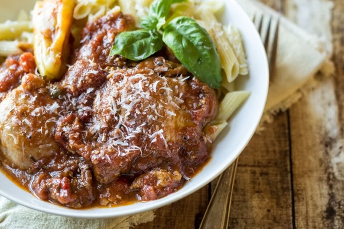 Chicken Cacciatore in a bowl, garnished with basil, with penne pasta and a pepperoncini on the side.