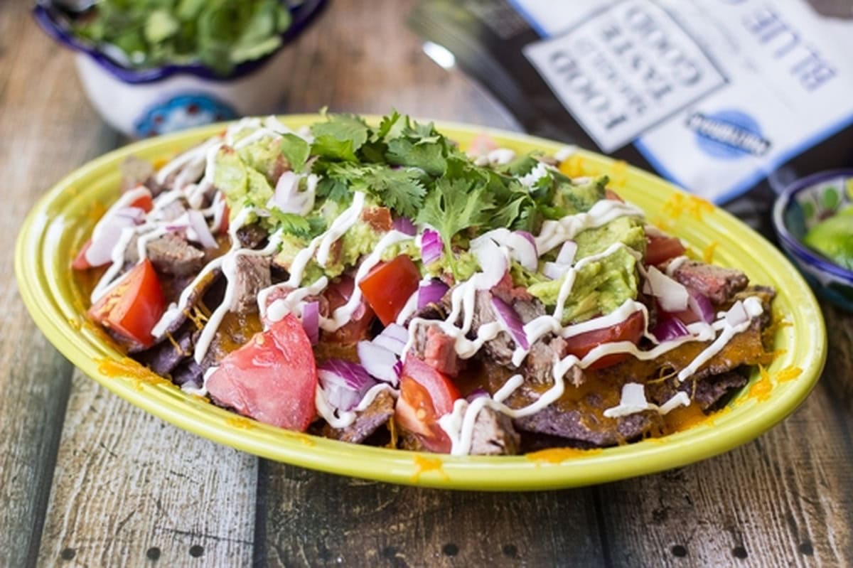 Carne asada nachos served on a plate alongside lime wedges and cilantro in small bowls.