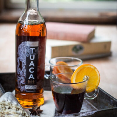 A bottle of Tuaca liqueur beside a glass of mulled wine garnished with an orange slice.