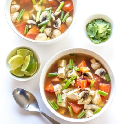 Two bowls of spicy Thai chicken soup and a bowl of lime wedges and cilantro leaves.