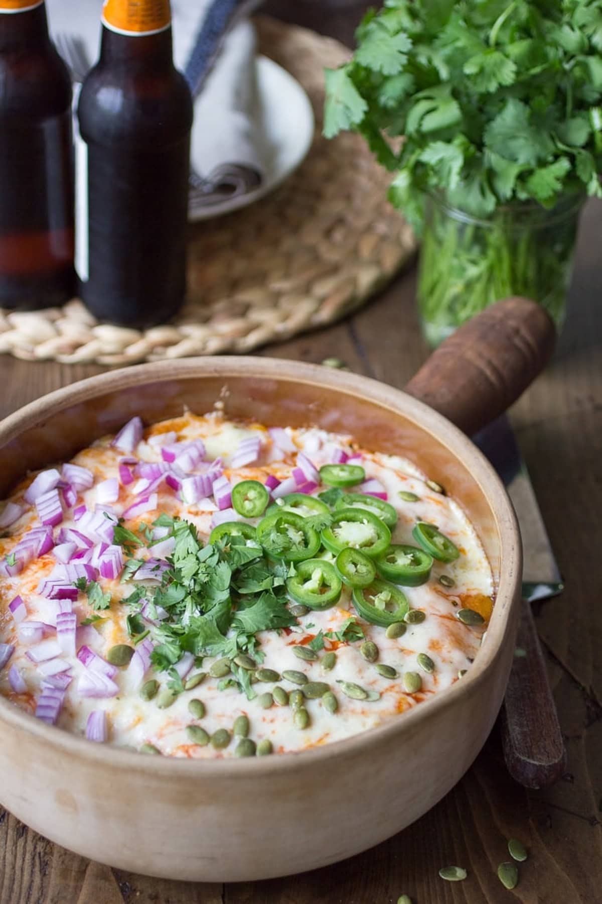 A wooden bowl filled with vegetarian enchilada pie, garnished with fresh jalape&ntilde;os, red onions, and cilantro.