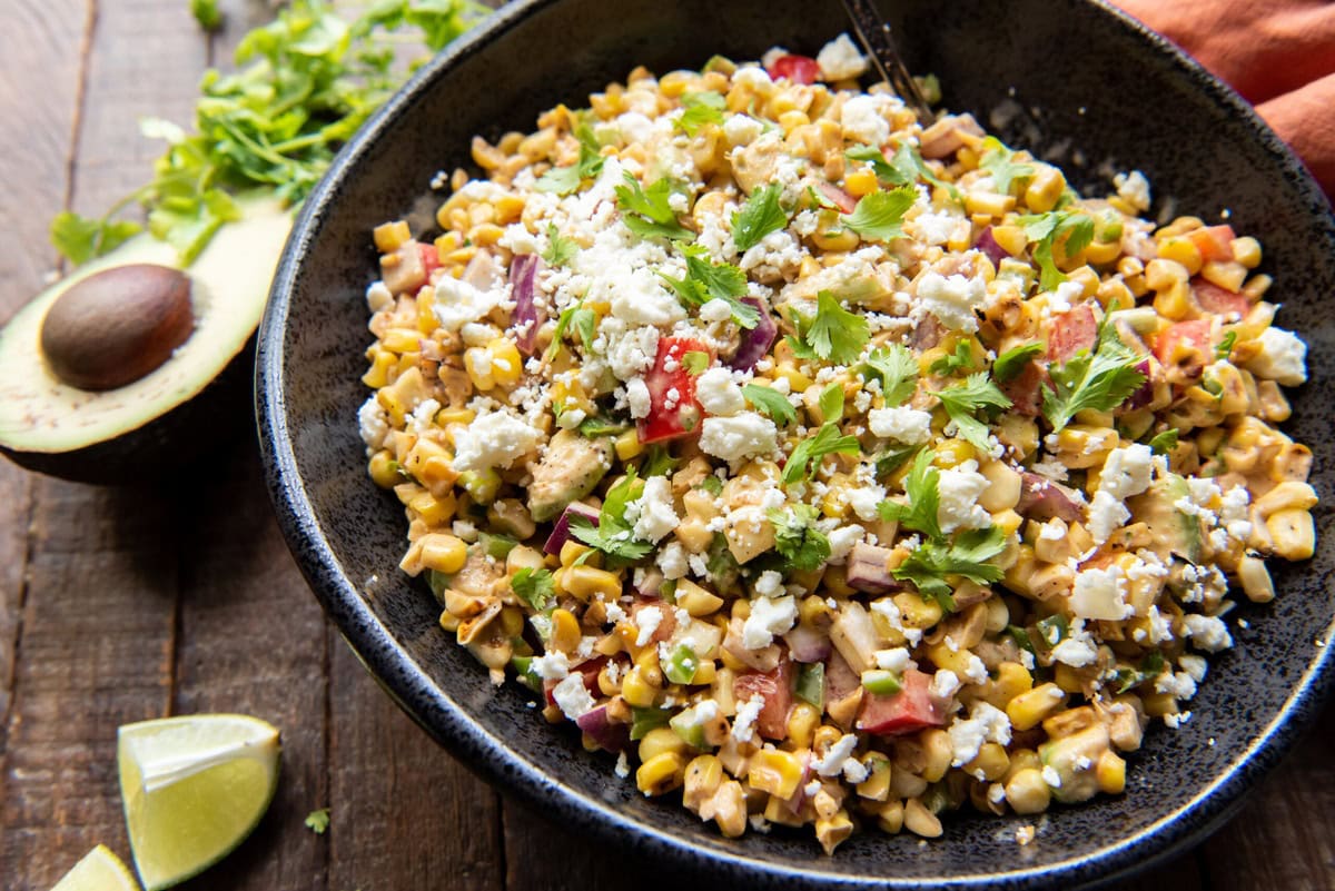 A bowl of Mexican corn salad, with avocado and lime on the side.